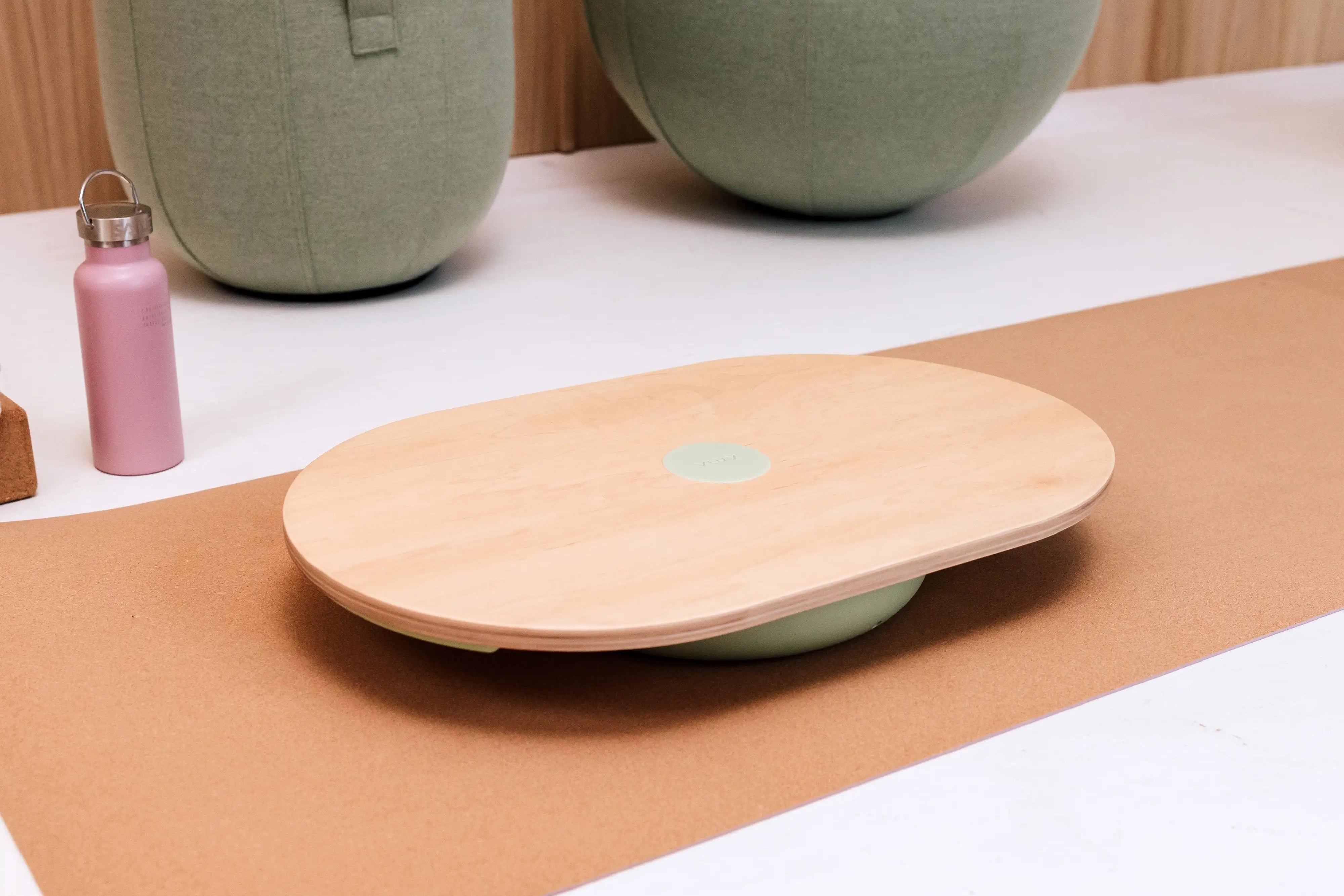 Holzbrett auf einem Yoga-Matte, neben grünen Sitzbällen und rosa Flasche.